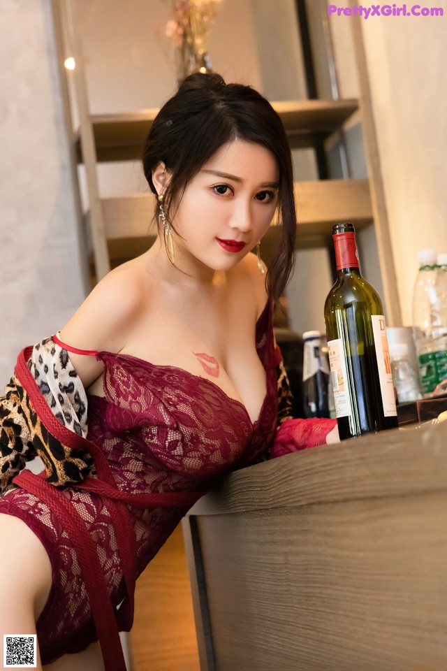 A woman in a red lingerie sitting at a table with a bottle of wine.