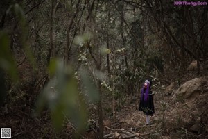 A woman with purple hair is sitting on a rock in the woods.