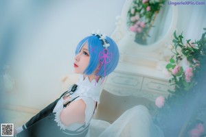 A woman with blue hair sitting on a bed with a teddy bear.