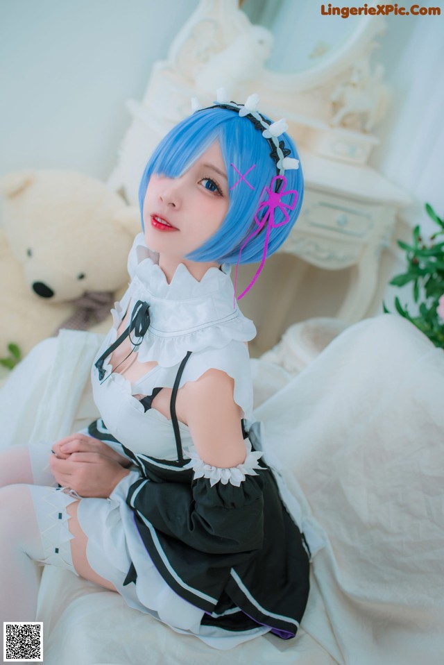 A woman with blue hair sitting on a bed with a teddy bear.
