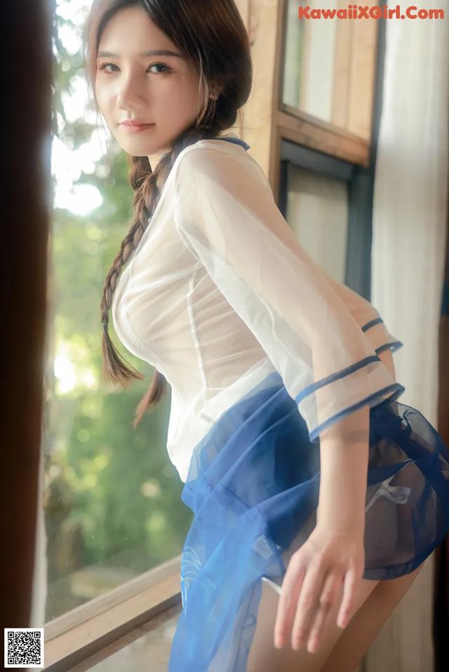 A woman in a white shirt and blue skirt posing by a window.